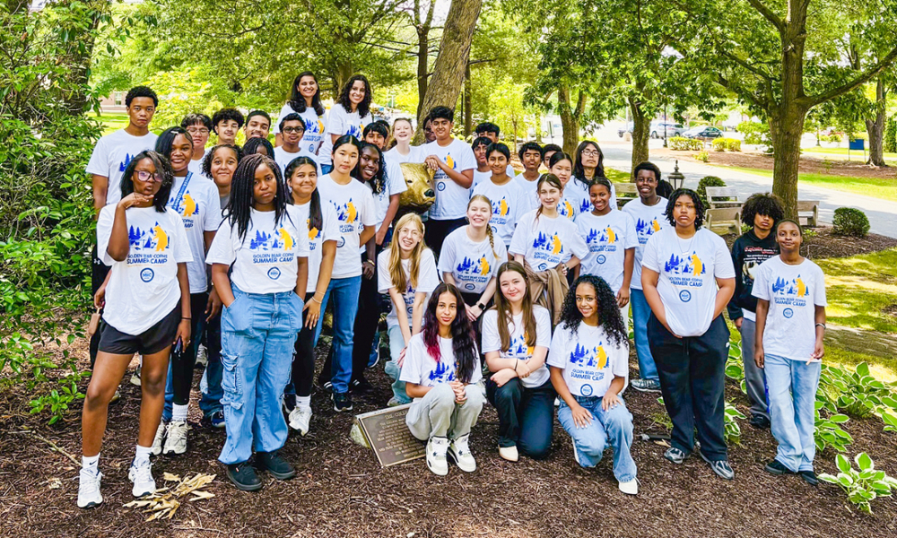 Approximately 45 students in Golden Bear Summer Camp t-shirts pose around WNE's Golden Bear.